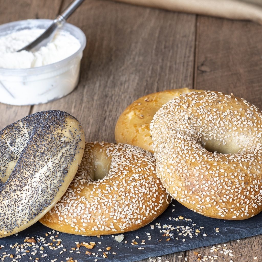 one plain bagel, two sesame seed bagels, and one poppy seed bagel sit on a black tray on a wood counter with a jar of cream cheese spread behind