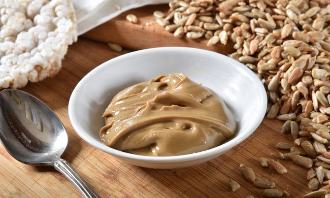 a ceramic bowl of sunflower butter with a silver spoon, a rice cake, and a pile of sunflower seeds next to it