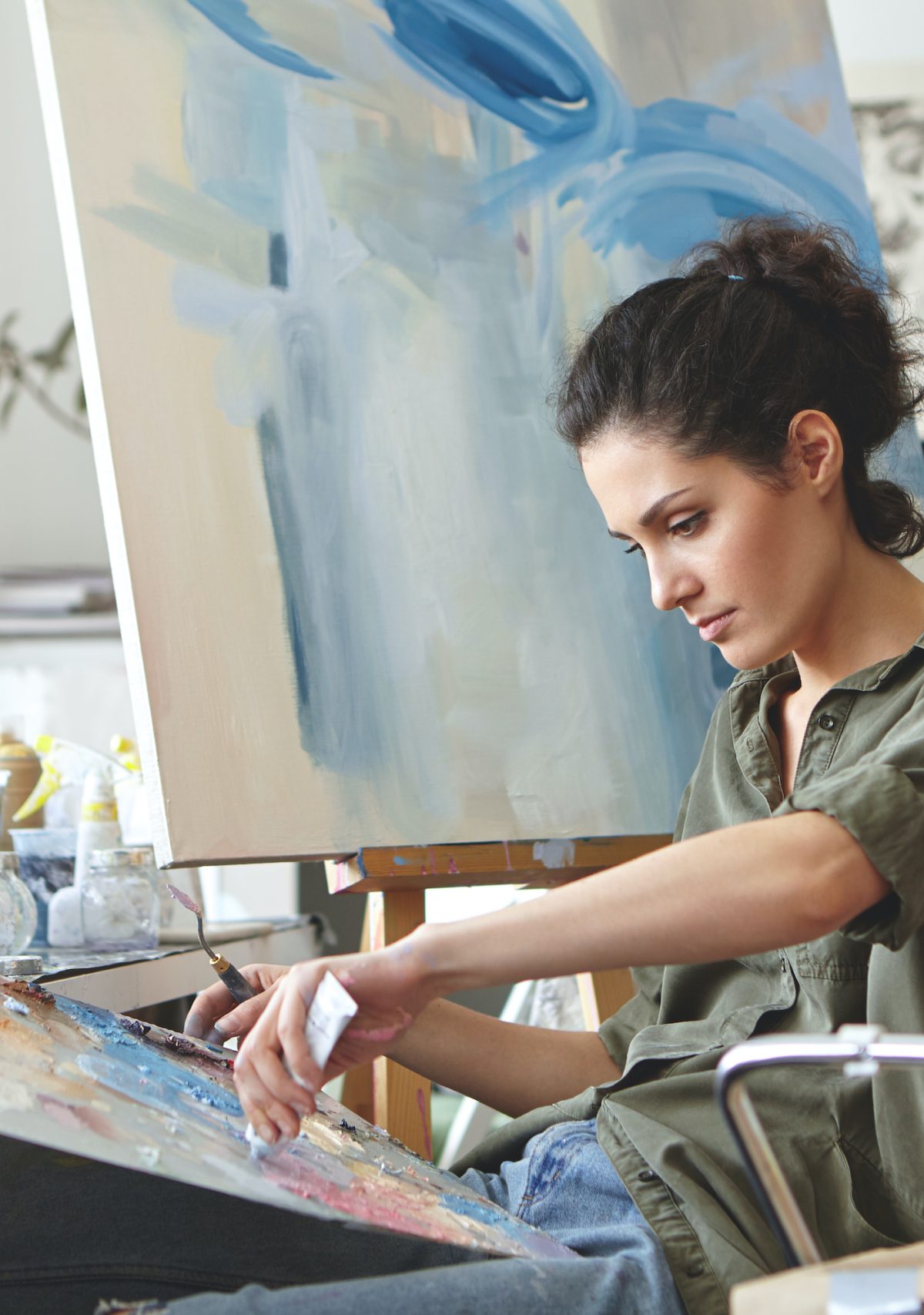 a woman with dark curly hair sits in front of a canvas and paints, surrounded by brushes in jars and paint bottles