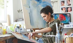 a woman with dark curly hair sits in front of a canvas and paints, surrounded by brushes in jars and paint bottles