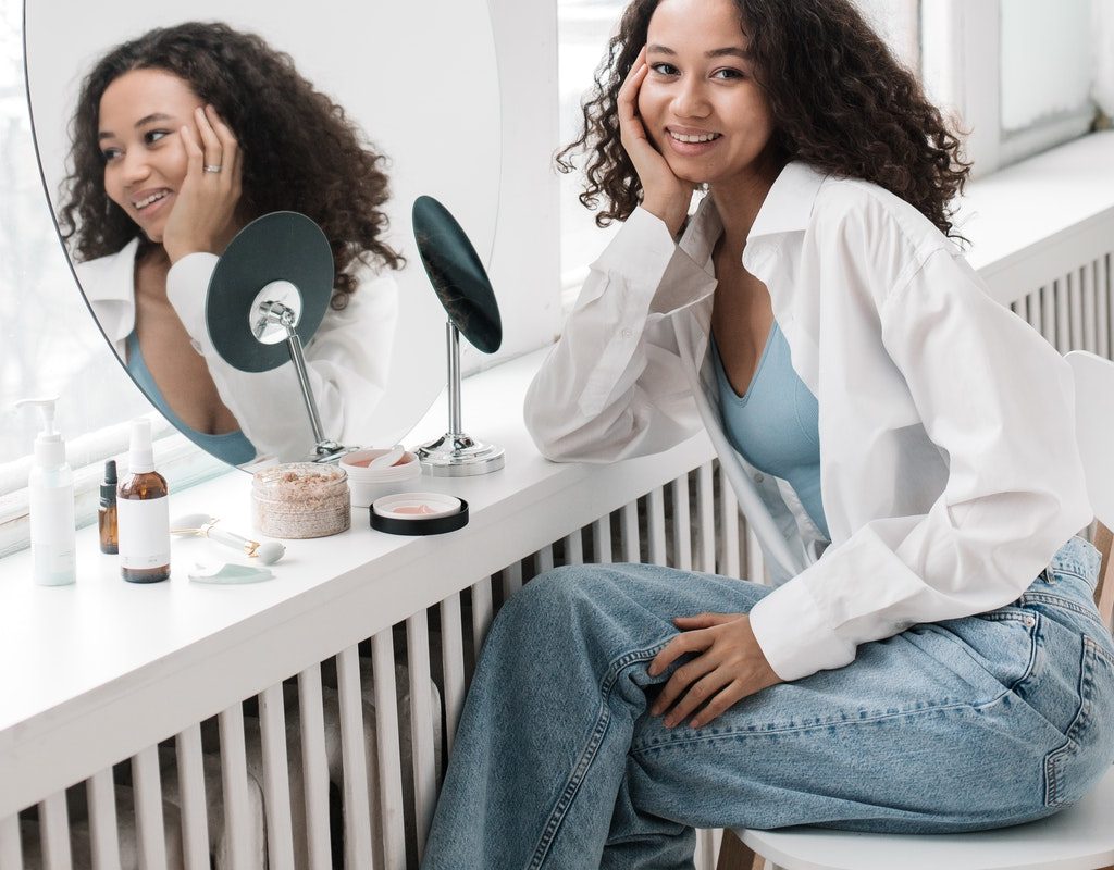 A woman smiling while sitting in front of a vanity with various skin care products on it.