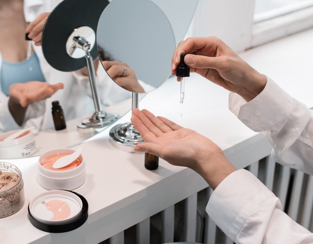 A woman dropping serum into one of her hands in front of a mirror where other beauty products are laid out.