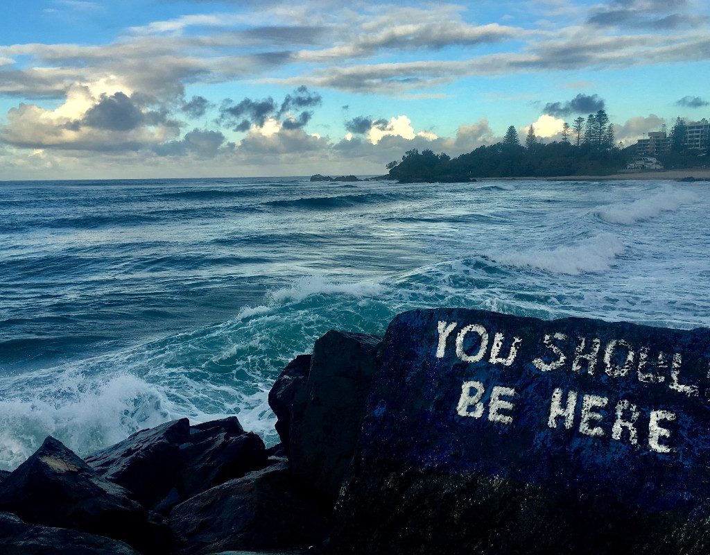 "You should be here" written in white lettering on a dark gray rock on a beach