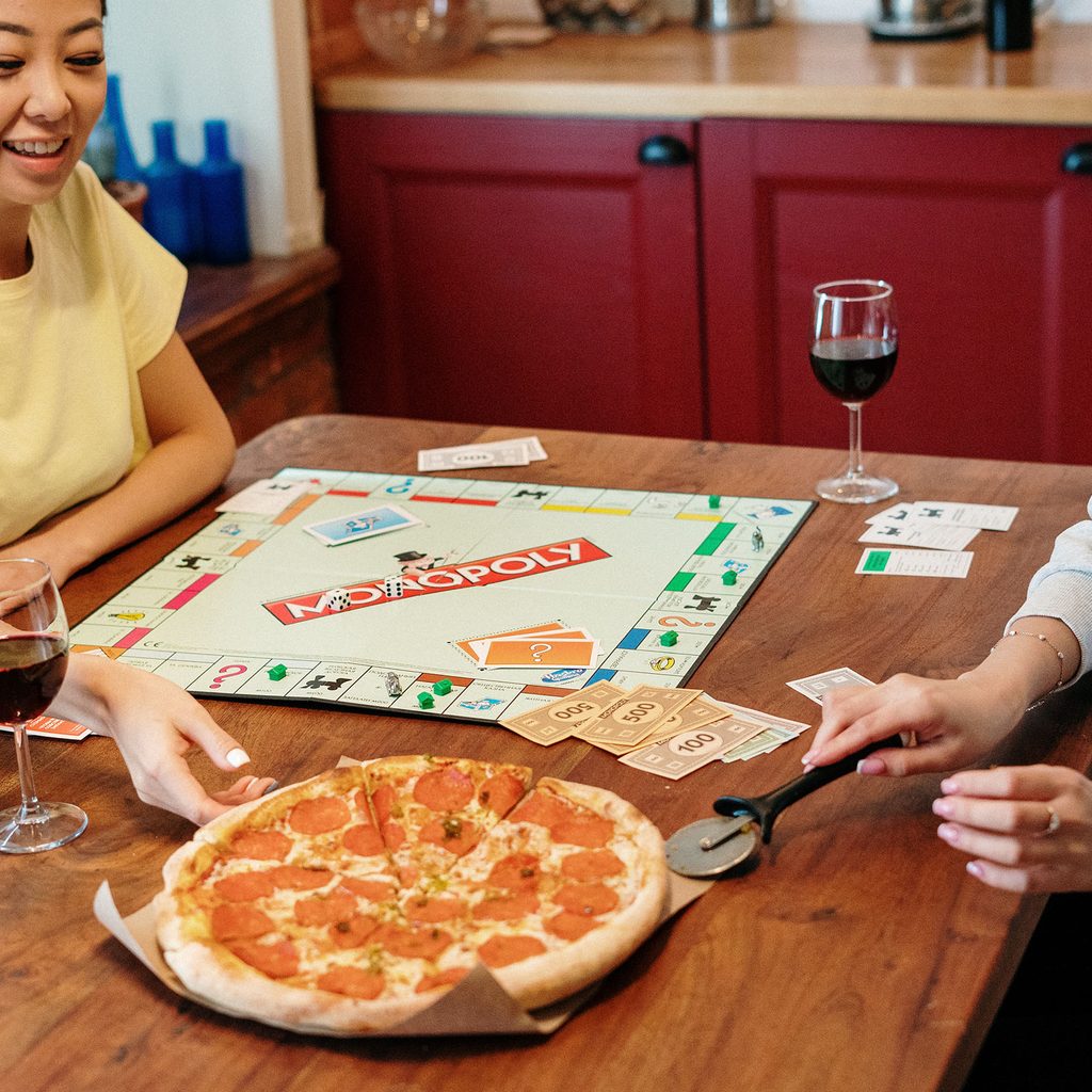 Two women playing Monopoly, eating pizza, and drinking wine