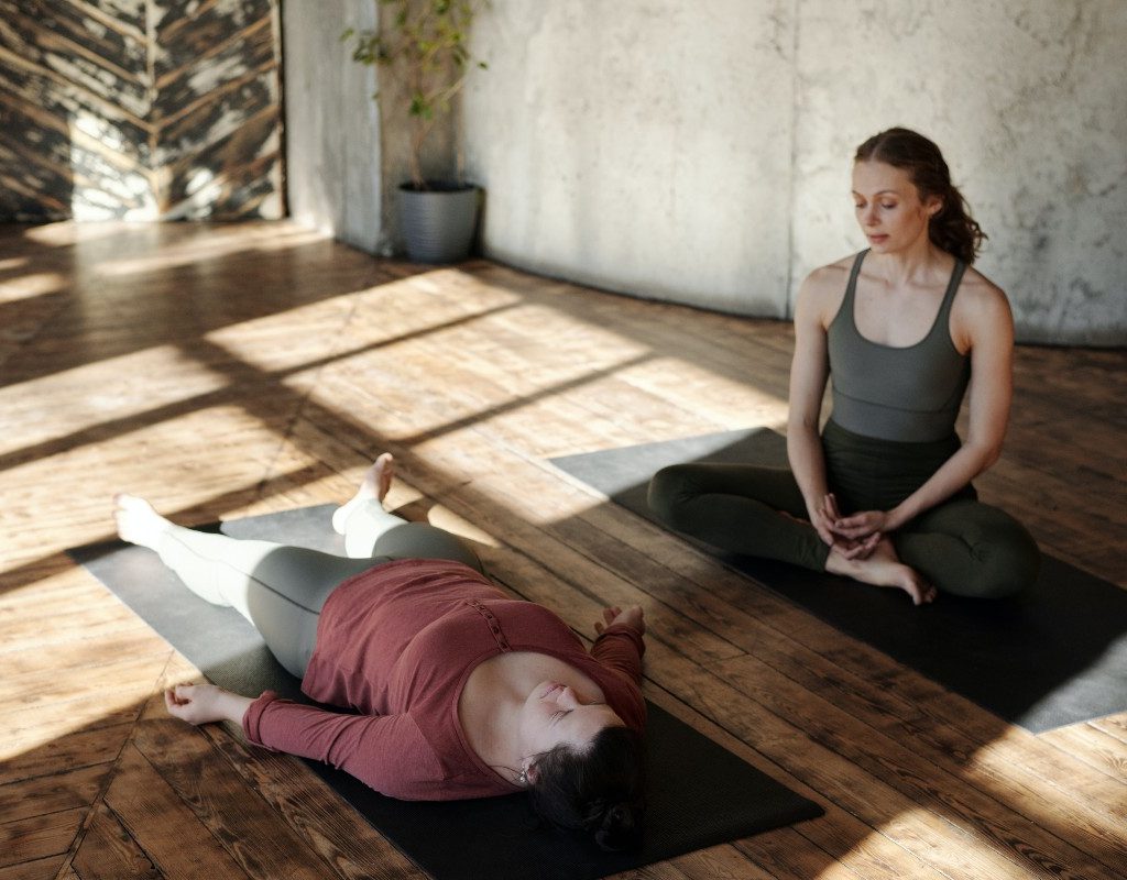 two women practicing yoga. one is in seated pose. the other is in savasana.