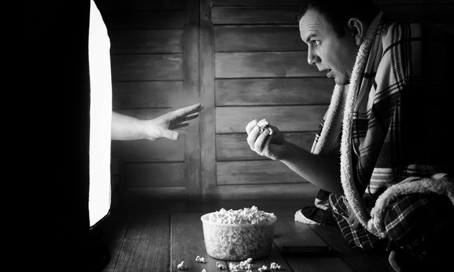A black and white image of someone watching a horror movie with a hand coming out of the tv to get him.