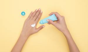A woman applying sunscreen from a tube with a yellow background.