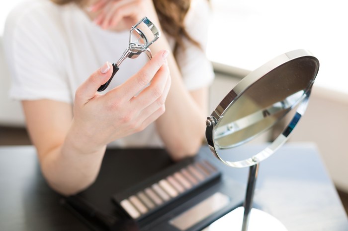 woman applying makeup and curling eyelashes in a mirror