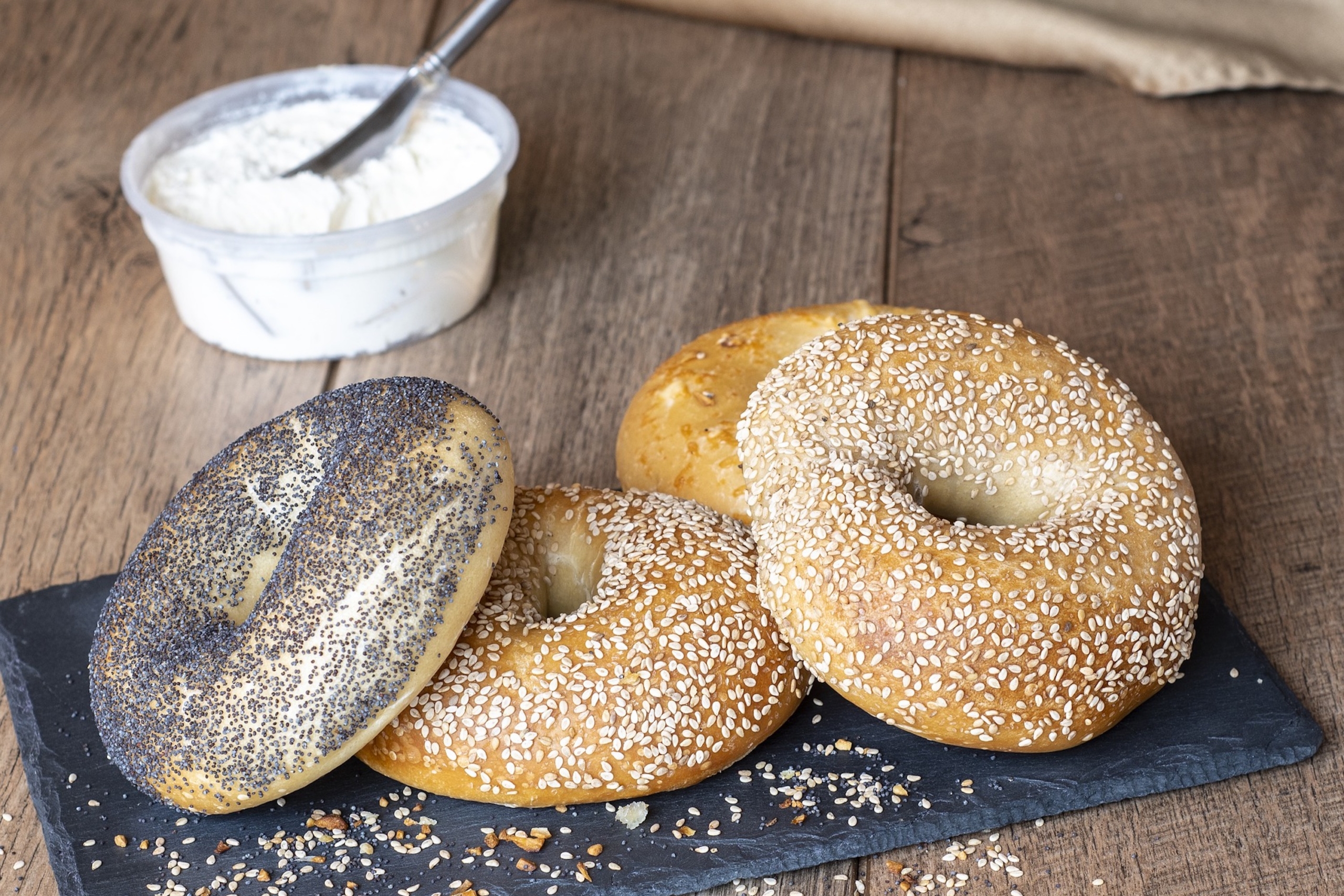 one plain bagel, two sesame seed bagels, and one poppy seed bagel sit on a black tray on a wood counter with a jar of cream cheese spread behind