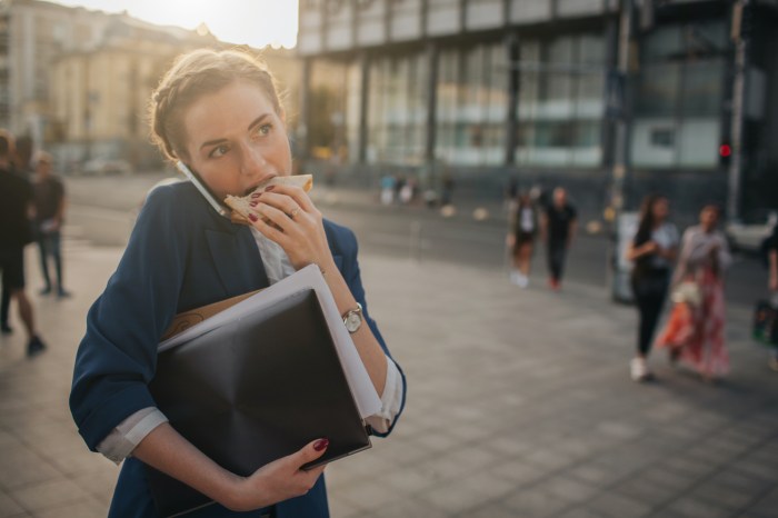 a businesswoman carries papers and folders in one arm, supports a cell phone to her ear with her shoulder, and eats a sandwich with the other hand