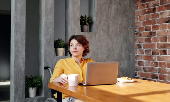 a woman sits at a table in front of a laptop, holding a cup of coffee, looking out to the distance