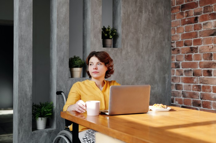 a woman sits at a table in front of a laptop, holding a cup of coffee, looking out to the distance