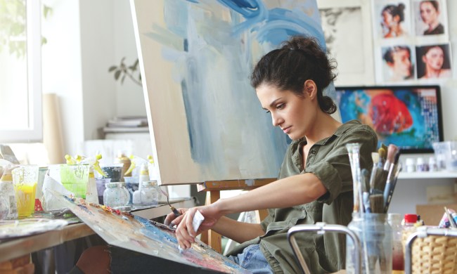 a woman with dark curly hair sits in front of a canvas and paints, surrounded by brushes in jars and paint bottles
