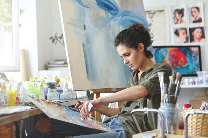 a woman with dark curly hair sits in front of a canvas and paints, surrounded by brushes in jars and paint bottles