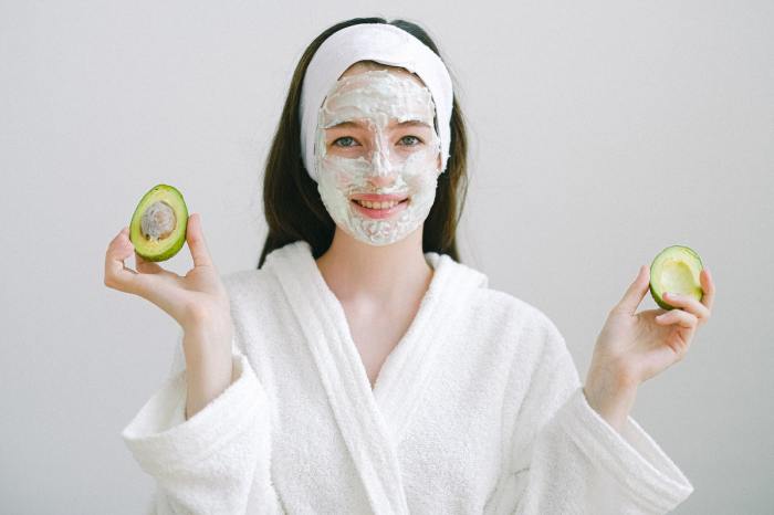 A woman with an exfoliating face mask one and holding half of an avocado in each hand.