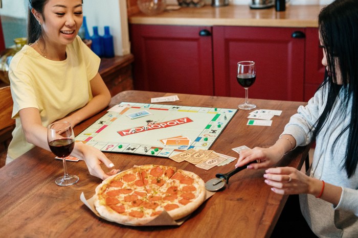 Two women playing Monopoly, eating pizza, and drinking wine