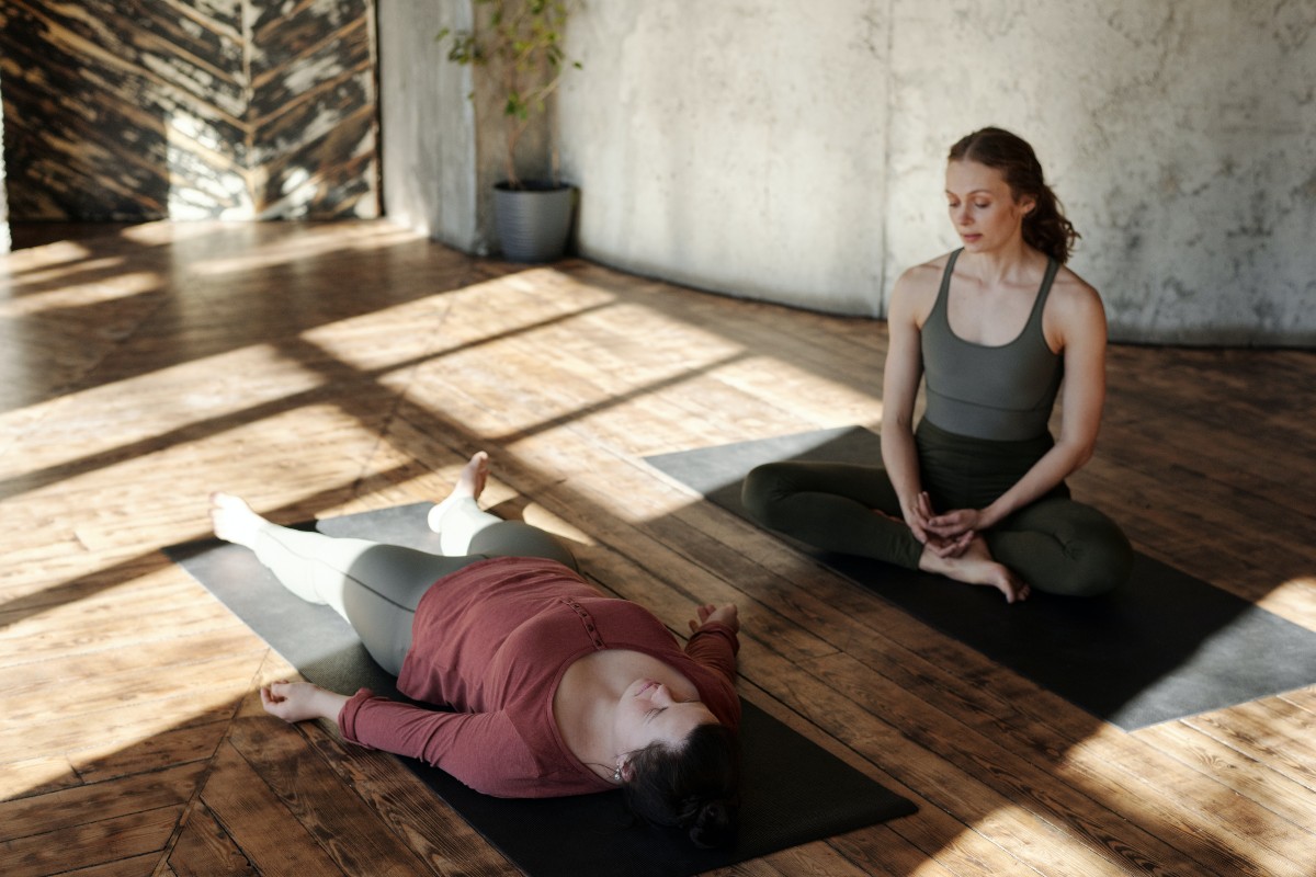 two women practicing yoga. one is in seated pose. the other is in savasana.