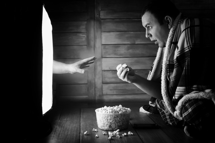 A black and white image of someone watching a horror movie with a hand coming out of the tv to get him.