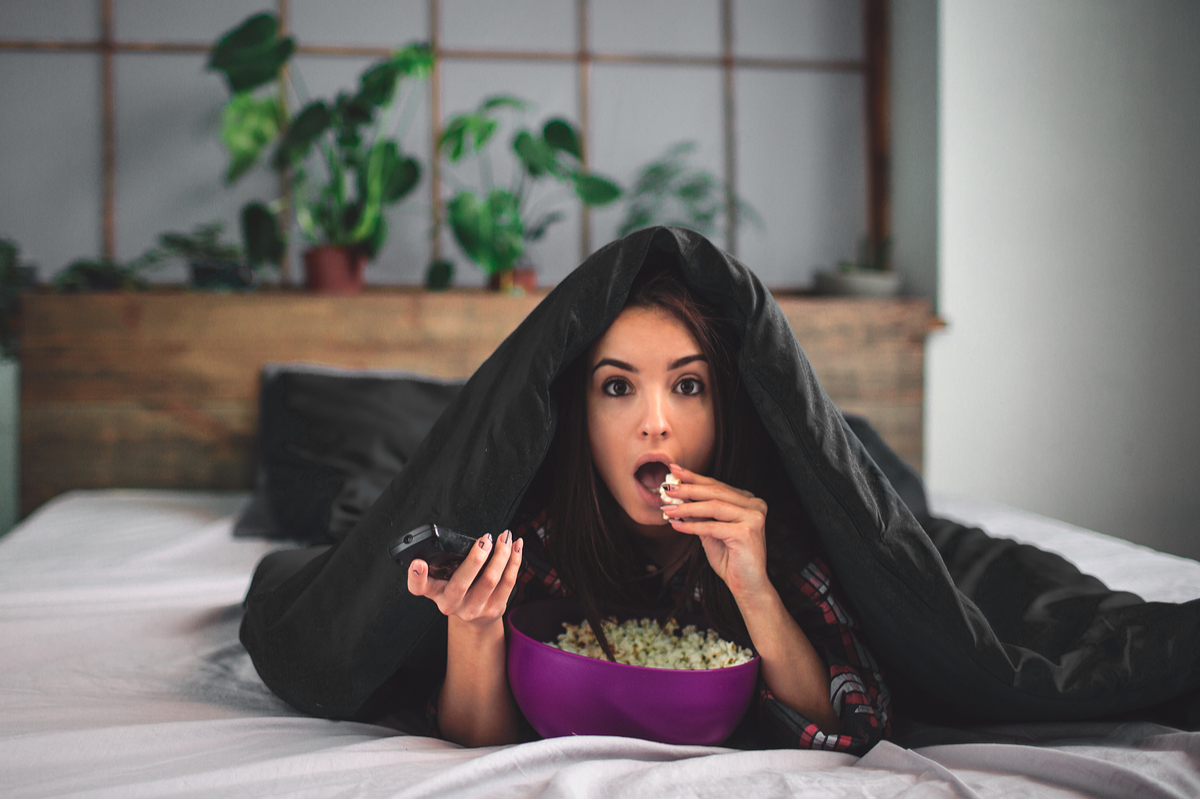 A woman watching a scary movie under the covers while eating popcorn.