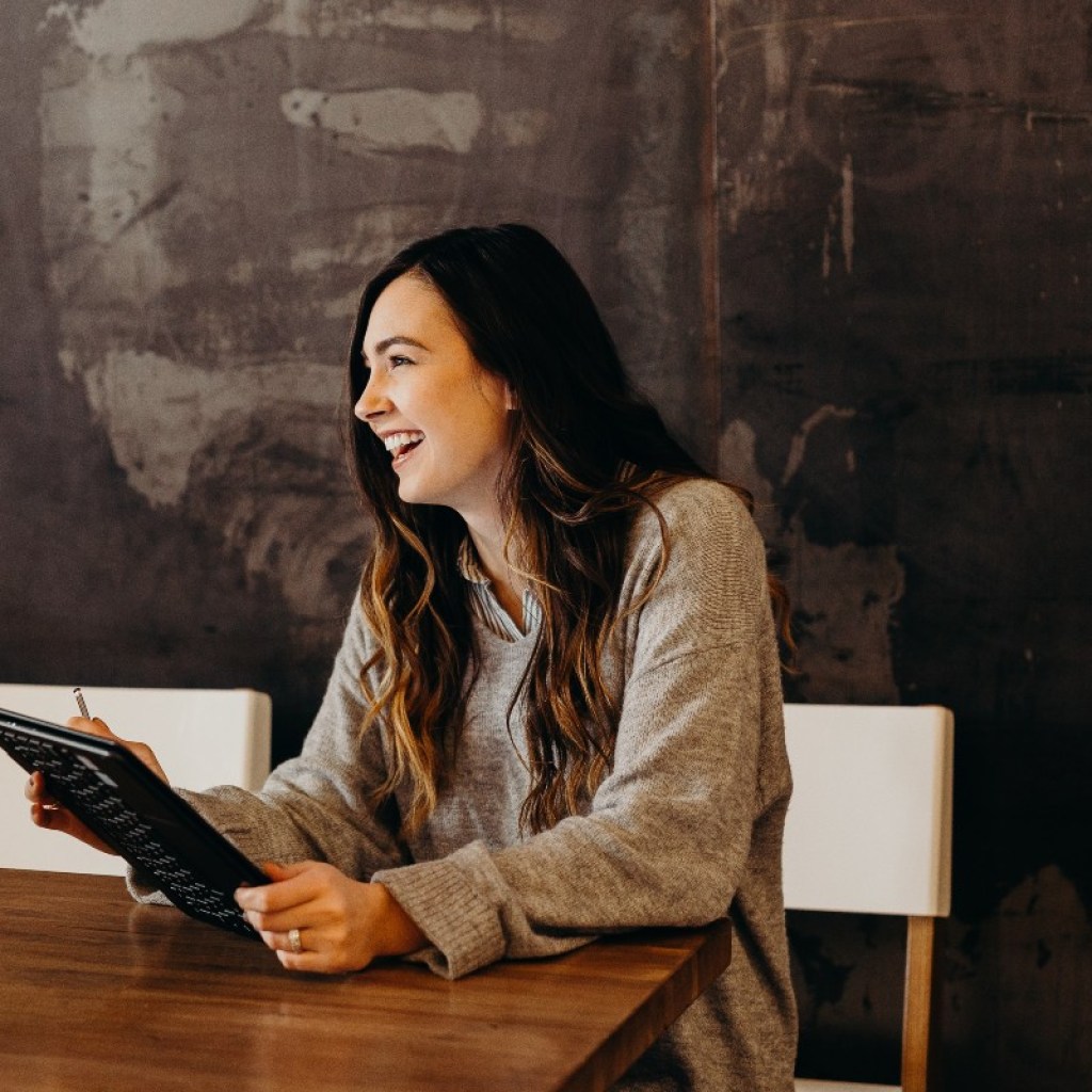 a woman sitting at a table holding a tablet and laughing