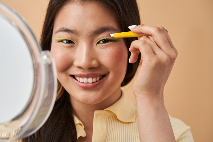 A woman with colorful eyeliner stands in front of a mirror holding tweezers to her eyebrow
