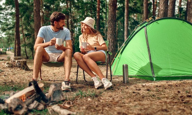 A young couple sit outside of their tent near a campfire, talking over coffee