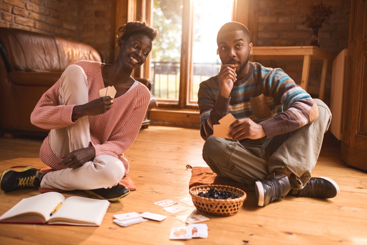 Young African American man playing cards with his girlfriend and thinking of his next move while woman is peeking at his cards.