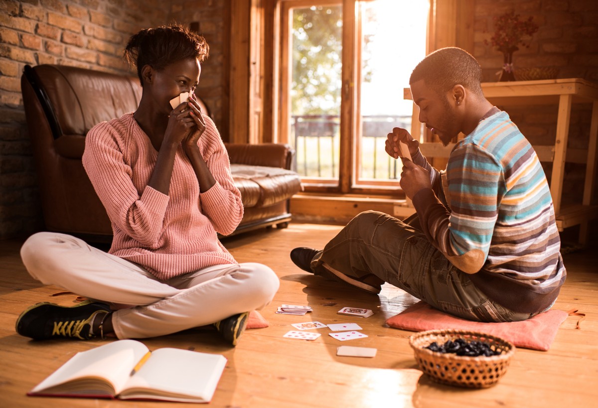 Young African American man playing cards with his girlfriend and thinking of his next move while woman is peeking at his cards.